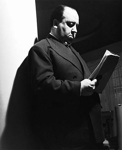 Movie dir. Alfred Hitchcock dourly reading script for the movie "Rebecca" as he stands against a pillar in his small apt. in the Wilshire Palms. Photo taken 1939 by Peter Stackpole